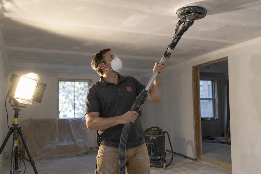 Crew member sanding a skim-coated ceiling with a pole sander under a raking work light