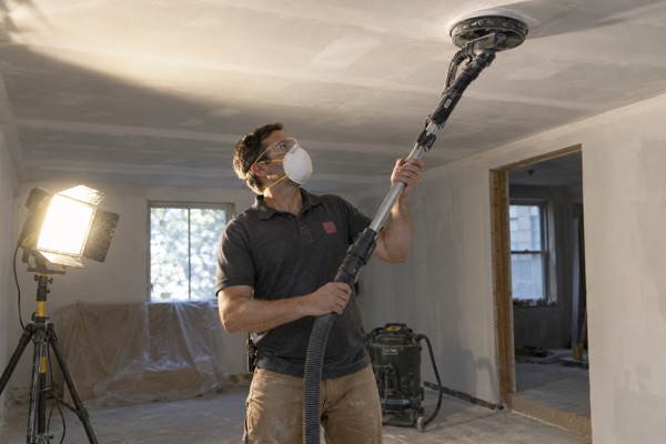 Crew member sanding a skim-coated ceiling with a pole sander under a raking work light