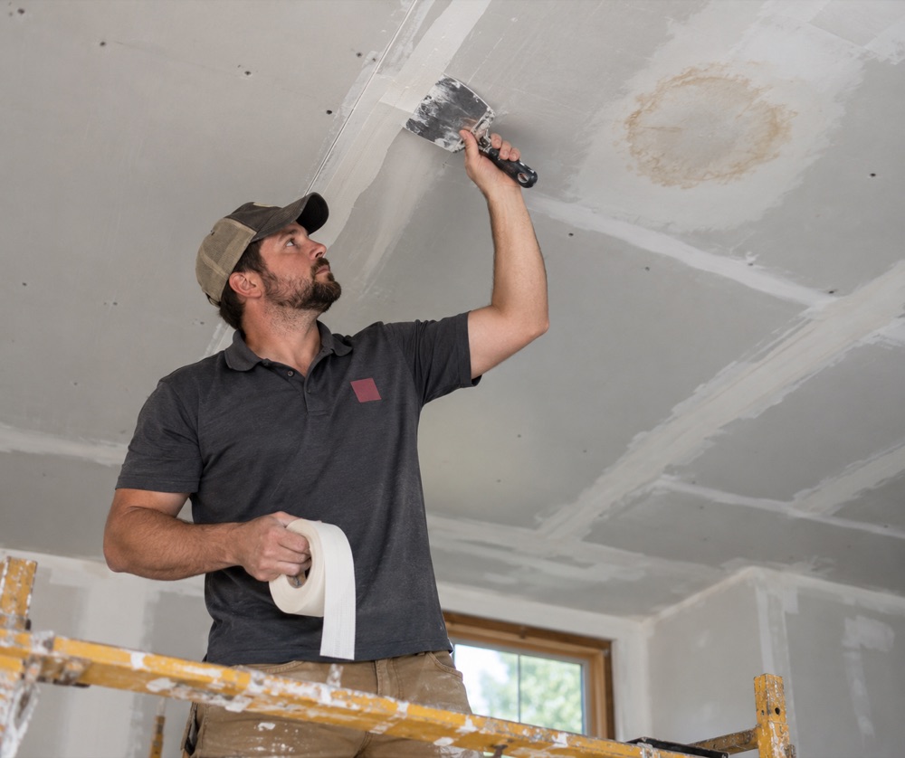 Crew member taping a drywall seam on a scraped ceiling near an old water stain